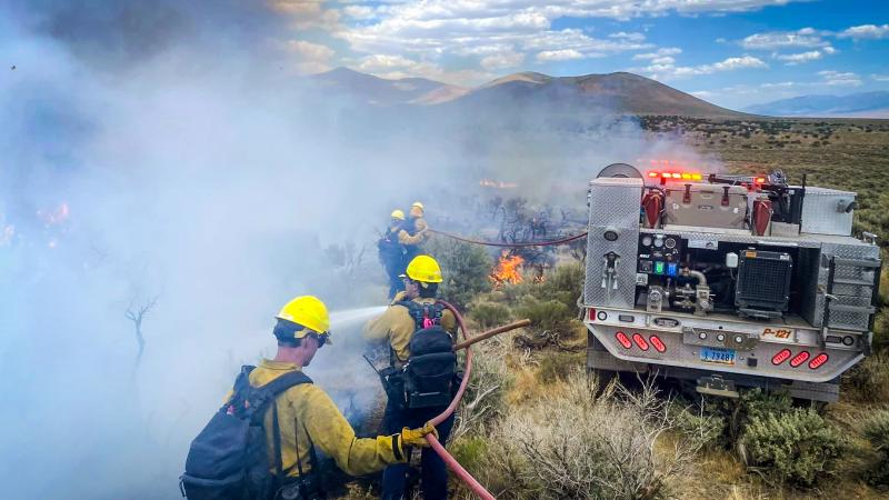 Firefighters walk alongside a vehicle driving away from the camera through dry shrubland. Small flames dot the landscape, and thick gray smoke obscures the landscape to the left of the firefighters. The firefighters use hoses attached to the vehicle to spray water onto the flames.