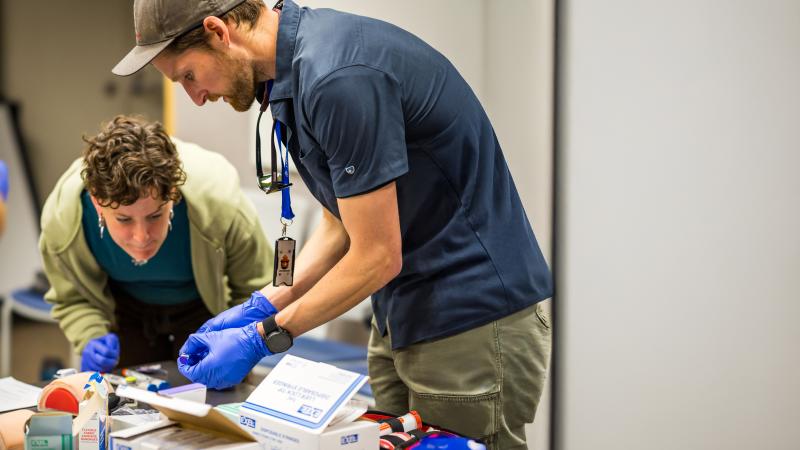 A man and a woman lean over a table scattered with medical supplies. The man holds a vial. The woman closely examines an item on the table. The table appears to be in an office.