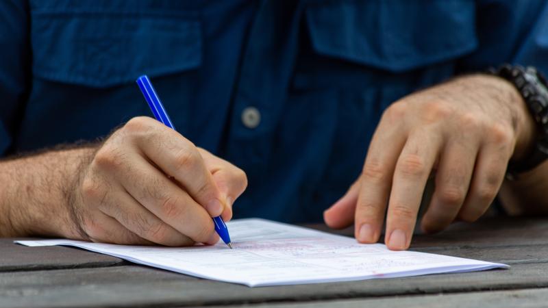 A close up shot of a man’s hands, one holding down the edge of a paper form while the other writes with a blue pen. He’s working at a weathered picnic table.