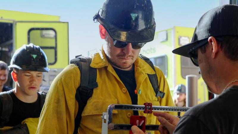 A wildland firefighter in a uniform and wearing a helmet and pack stands on a scale, looking down to watch the result as someone on the other side adjusts the scale. Another firefighter stands in line behind him. In the background, two wildland fire vehicles and another firefighter are visible. It’s a sunny, cloudless day.