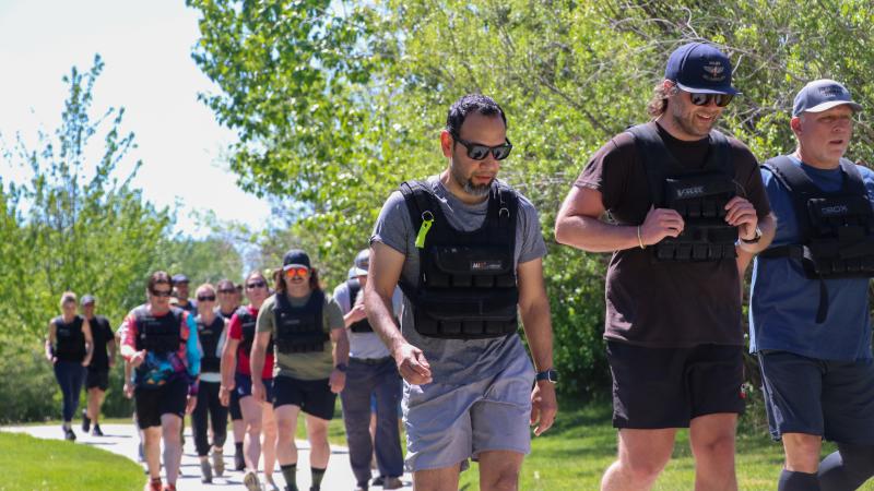 People wearing athletic shorts, T-shirts, and sneakers walk and jog along a wide paved path, with three people ahead of a larger group while two others jog in the back. They are all wearing black weighted vests. They are also wearing a mix of baseball hats and sunglasses. The path is lined by vibrant green grass and, on one side, verdant trees. It’s a bright day with a cloudless sky.