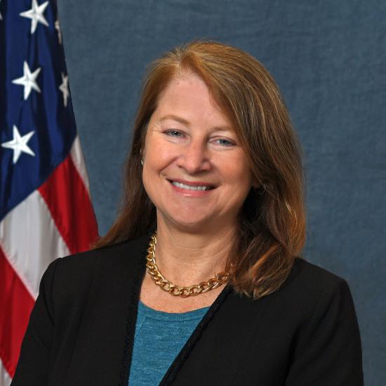 Headshot of woman in blue shirt and black blazer in front of American flag