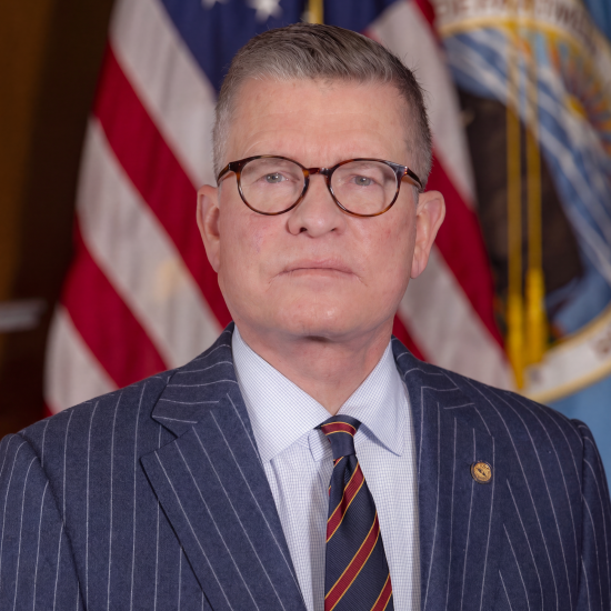Portrait of Kevin Lilly in navy striped suit and dark rimmed glasses in front of American Flag and DOI flag. 