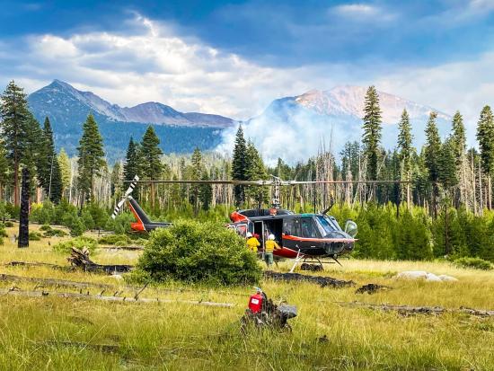 Members of a Yosemite helitack crew work around a helicopter in a grassy field. Trees border the field, with smoke rising from their midst. Mountains rise in the distance under a stormy sky. Photo by Yosemite National Park.
