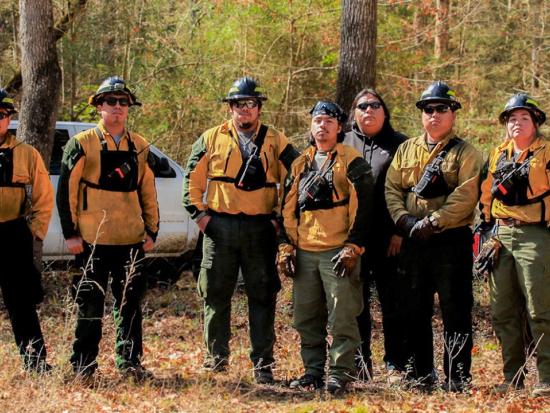 Members of the Alabama-Coushatta Tribe fire management team stand in a leaf-strewn clearing staring at the camera with determined expressions. Behind them is a dense, mixed stand of trees and two vehicles. Photo by Claire Everett, The Nature Conservancy.