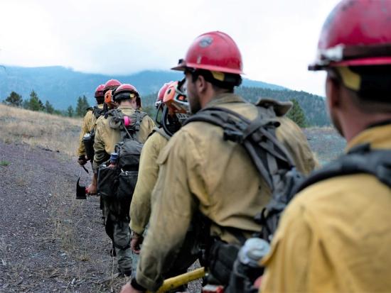 Wildland firefighters hike in a line wearing their gear and carrying tools. 