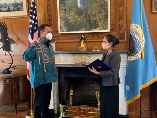 Secretary Deb Haaland swears in Bryan Newland in front of two flags and a fireplace. 