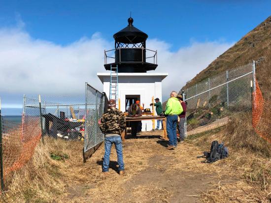 Workers completing repairs on the Punta Gorda Lighthouse, surrounded by metal fencing with a blue sky and ocean in the background.