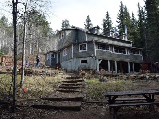 Two-story wood shingle lodge building on raised piers with an enclosed porch, located in a wooded setting with stone terracing and log fencing. 