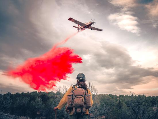 A wildland firefighter watches as a single engine air tanker drops red retardant on a wildfire. The firefighter's back is to the viewer as the plane flies overhead, with smoke and clouds in the background. BLM photo. 