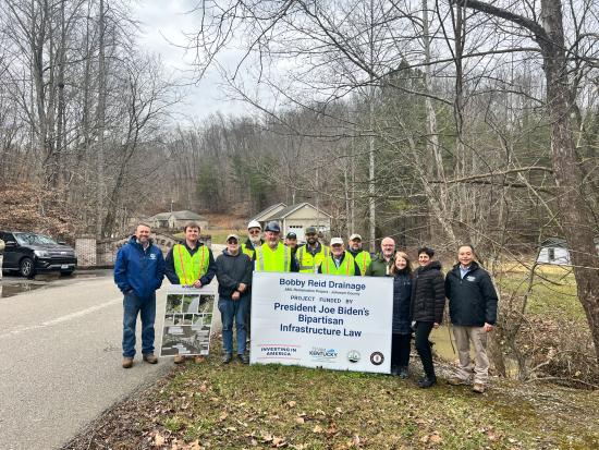 Group of people in safety gear standing behind Bipartisan Infrastructure Law Sign
