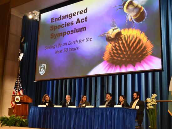 6 people sitting on stage with a presentation slide "Endangered Species Act Symposium"