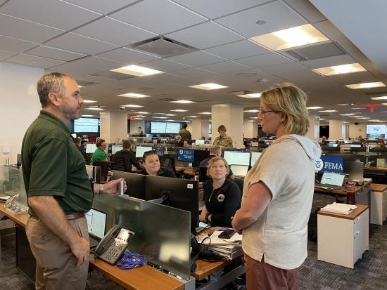 A man and woman stand in a conference room filled with computers and other personnel. 