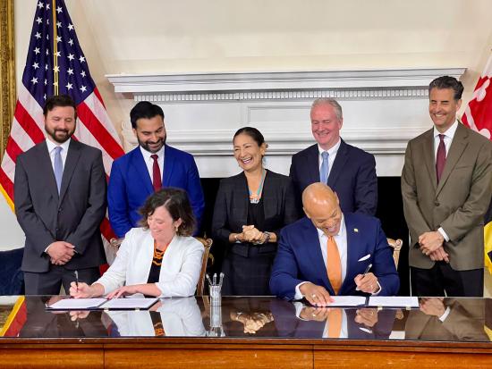 Group of Department Leadership standing behind table. Two people are seated and signing documents.