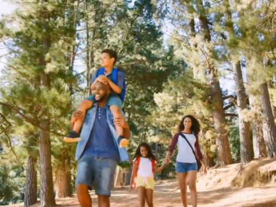 A smiling family walks along a national park forest path, the son on his father’s shoulders, the mother holding her daughter’s hand.