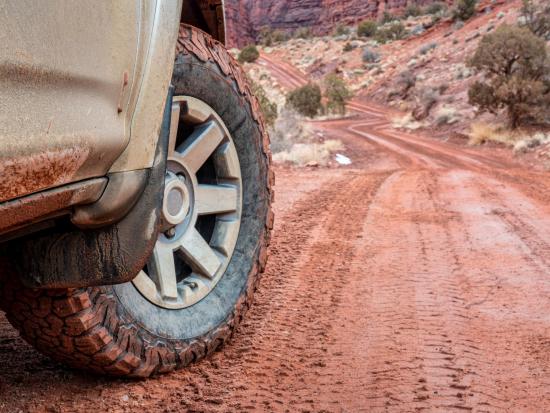 A SUV is on a dirt road. You can see the front tire of the vehicle and the red dirt road in front of it. 