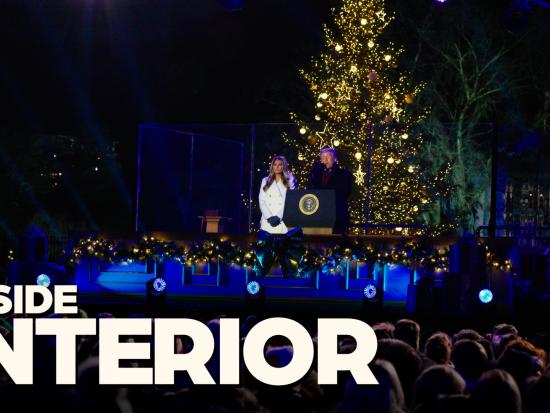 President Trump and the First Lady stand behing a podium at night and speak to an audience as the National Christmas Tree is lit in white behind them