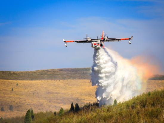 A small airplane flies low to the ground at an angle toward the camera. Water pours out of open doors at the bottom of the plane, billowing down to the ground. The spray at the back catches the light and glows orange, yellow, and a hint of turquoise. The water is dropped on yellowish-green vegetation dotted with low trees and a faint hint of smoke. In the background, yellow-brown hills stretch across the horizon under a blue sky.