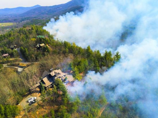 A large house with two trucks parked outside is seen from above. A handful of other, smaller homes are scattered through the surrounding forested area, connected by narrow, winding roads. White smoke rises in a wedge formation from the trees to the right of the homes, obscuring the forest from view. To the left, the forested hillside descends toward a grassy plain, with more forested mountains visible in the distance.
