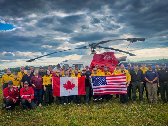 A group of people in different yellow, red, black, and blue uniforms pose for a photo in the middle of a grassy field. Those in the center hold up an American flag and a Canadian flag. A helicopter is parked immediately behind them. A row of parked cars is just visible at the edge of the field to the left. Int eh background, dense trees line the far edge of the field. The sky is a moody patchwork of dark clouds, blue sky, and light clouds illuminated in gold by the setting sun.