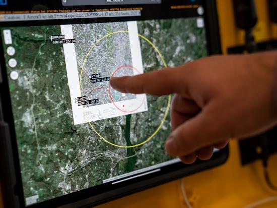  A hand extends toward a tablet screen mounted on a yellow panel, a finger pointing toward highlighted text and a red circle near the center of the screen. The screen displays a detailed topographical map with overlaid concentric circles and three small airplane icons with associated text. A portion of the map is magnified in an overlaid callout box.