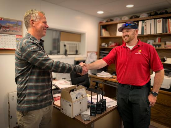 Two men in casual attire shake hands, smiling at each other. Under their clasped hands is a row of radios and a box with more equipment sitting on the corner of a desk. They are standing in a small office. The desk is scattered with papers. The shelves along the back wall hold binders and supplies. On the wall to the left, a calendar displays a photo of a red fire truck.