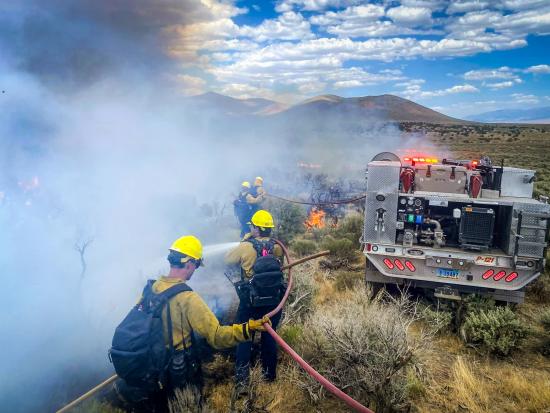 Firefighters walk alongside a vehicle driving away from the camera through dry shrubland. Small flames dot the landscape, and thick gray smoke obscures the landscape to the left of the firefighters. The firefighters use hoses attached to the vehicle to spray water onto the flames.