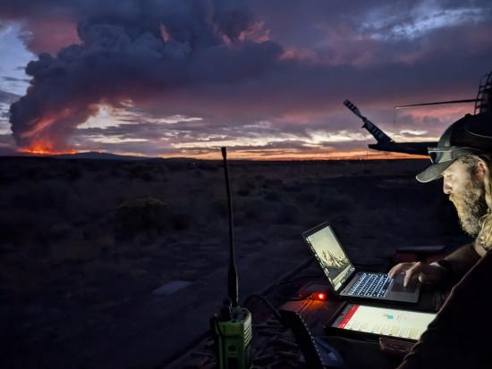 Two people work on a laptop and a tablet at dusk, standing at a makeshift table set up in a field of sagebrush. A radio and other equipment clutter the table. Their faces are illuminated by the glow of the screens. The tail of a helicopter is visible behind them. An orange-red fire glows on the mountains in the distance, a plume of smoke rising into the sky and merging with the scattered clouds that are lit by the last light of the sunset.