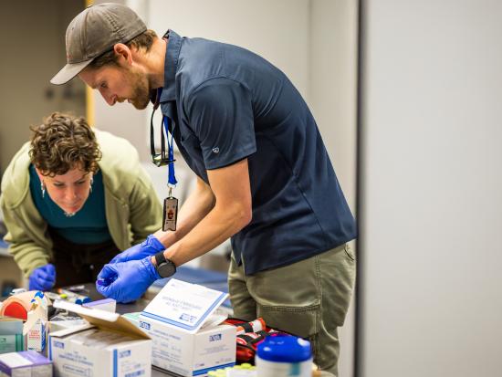 A man and a woman lean over a table scattered with medical supplies. The man holds a vial. The woman closely examines an item on the table. The table appears to be in an office.