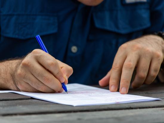 A close up shot of a man’s hands, one holding down the edge of a paper form while the other writes with a blue pen. He’s working at a weathered picnic table.