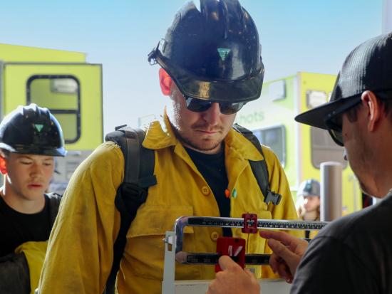 A wildland firefighter in a uniform and wearing a helmet and pack stands on a scale, looking down to watch the result as someone on the other side adjusts the scale. Another firefighter stands in line behind him. In the background, two wildland fire vehicles and another firefighter are visible. It’s a sunny, cloudless day.