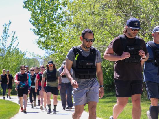 People wearing athletic shorts, T-shirts, and sneakers walk and jog along a wide paved path, with three people ahead of a larger group while two others jog in the back. They are all wearing black weighted vests. They are also wearing a mix of baseball hats and sunglasses. The path is lined by vibrant green grass and, on one side, verdant trees. It’s a bright day with a cloudless sky.