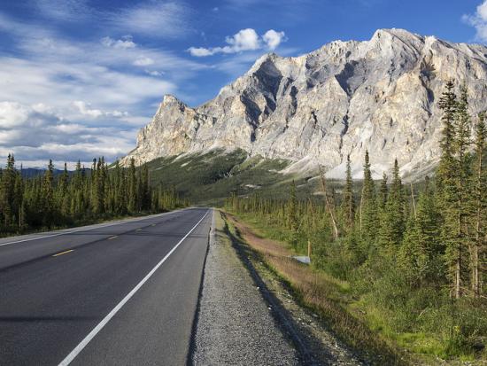 Highway stretching toward tall rocky mountains with pine trees lining both sides under a blue sky