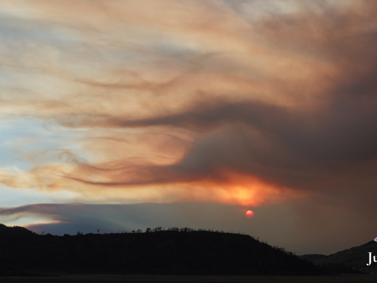 Orange-tinged wildfire smoke drifts across the sky as the sun, an orange ball glowing through the clouds, sets over silhoutted hills. A purpose ribbon appears in the lower right corner above the text "June 27, 2026."