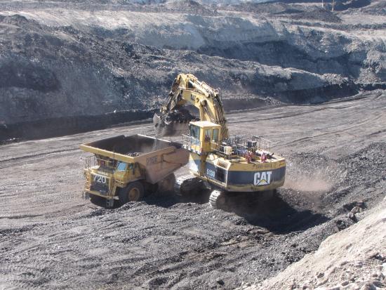 A CAT hydraulic excavator loading a yellow CAT haul truck with coal, set against tiered gray overburden slopes in an open-pit mine. 