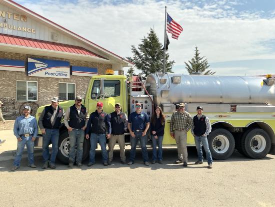 Nine people pose and smile in front of a large truck with a tank 