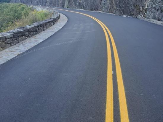 Freshly paved mountain road with double yellow lines curving beside a stone wall and steep, tree-covered rock face