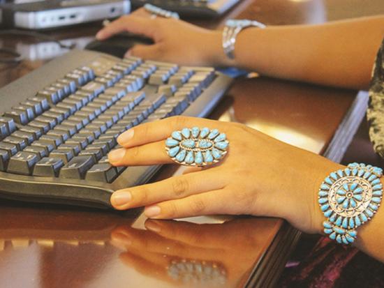 A view of a person's hands with turquoise jewelry typing at a keyboard and using a computer mouse.