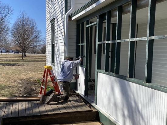 Man kneels to paint the side of a white house with green trim; small orange ladder stands next to him.