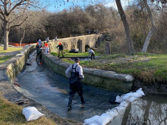 A group of eight construction workers repair a curved stone aqueduct surrounded by bare trees