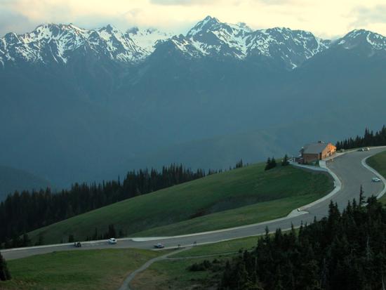 Winding road on grassy hillside with snow-capped mountains in the background. 