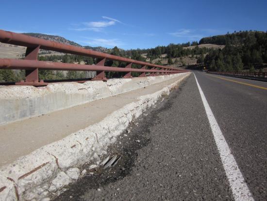 Close-up photo of a paved road with red railings with pine trees and hills in the distance.