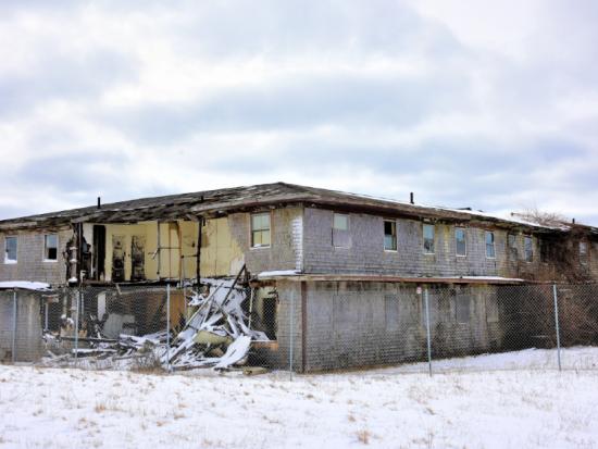 A large building sits behind fencing with walls torn away, exposing the interior of the building