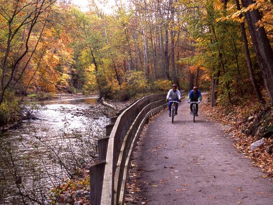 Two people on bicycles on a flat trail surrounded by fall forest colors. A wooden fence in the center divides the trail to the right and the river to the left.