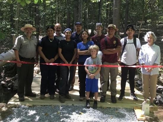 Twelve people standing on wooden bridge in the forest getting ready to cut the red ribbon to open the bridge to visitors.