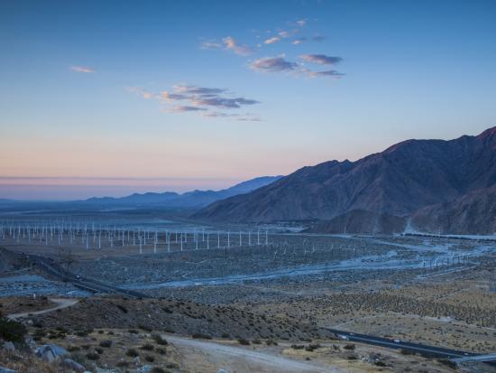 Many wind turbines stand tall in the desert with mountains in the distance