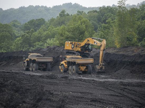 Yellow construction equipment amidst mine waste with green trees and hills in the background