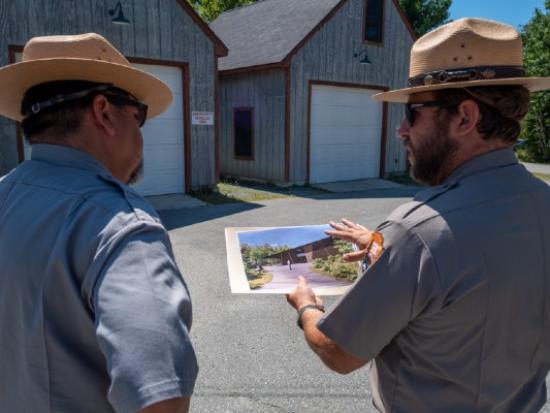 Two uniformed men stand in front of buildings, one holding an image of a building.