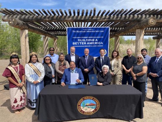Group of people standing around a table with the logo Gila River Indian Community, Arizona. There is a sign in the backdrop "Building a Better America"