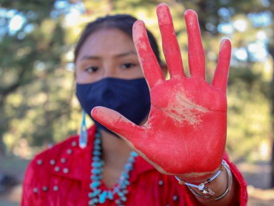 An Indigenous woman holds up her hand that has red paint on it and stands outside